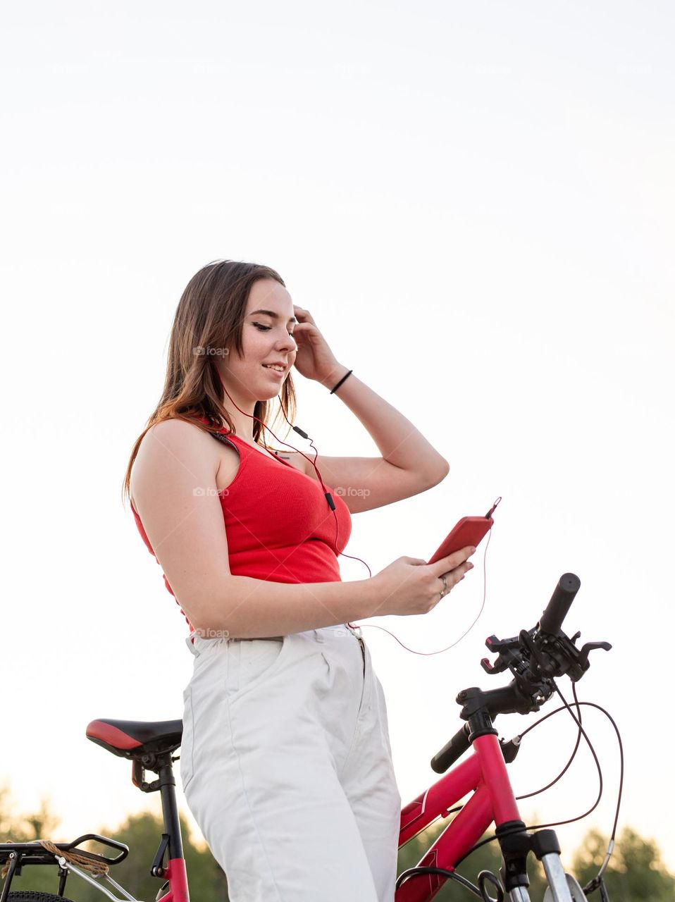 young smiling woman in red shirt and white pants listening to the music standing near her bicycle