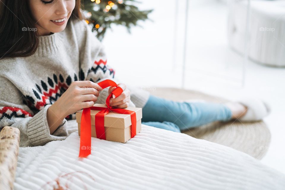 Young woman in cozy sweater with gift box with red ribbon in room with Christmas tree at home