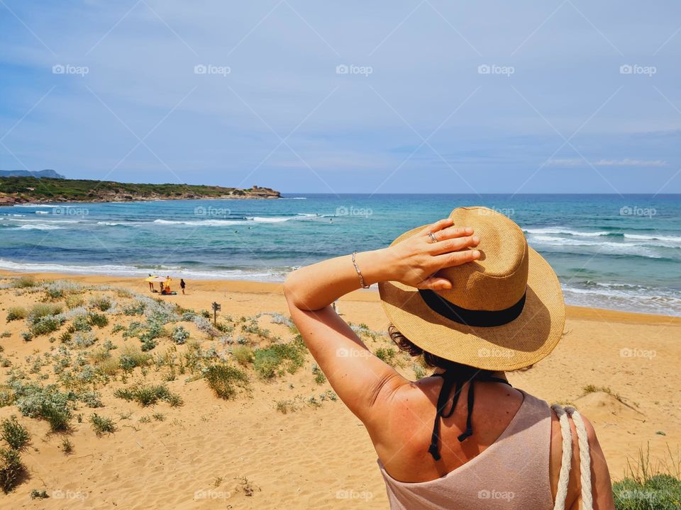 woman from behind with straw hat looks at the sea and the wild beach of Sardinia