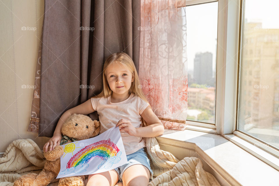 Happy little Caucasian girl with blonde hair painting rainbow during COVID-19 coronavirus quarantine and self isolation 