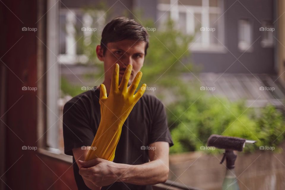 Portrait of one young handsome Caucasian brunette man standing by a window without frames and glass, looking into the sky and putting on yellow rubber gloves, getting ready for work on a summer evening, etc. from the side close up with selective focu