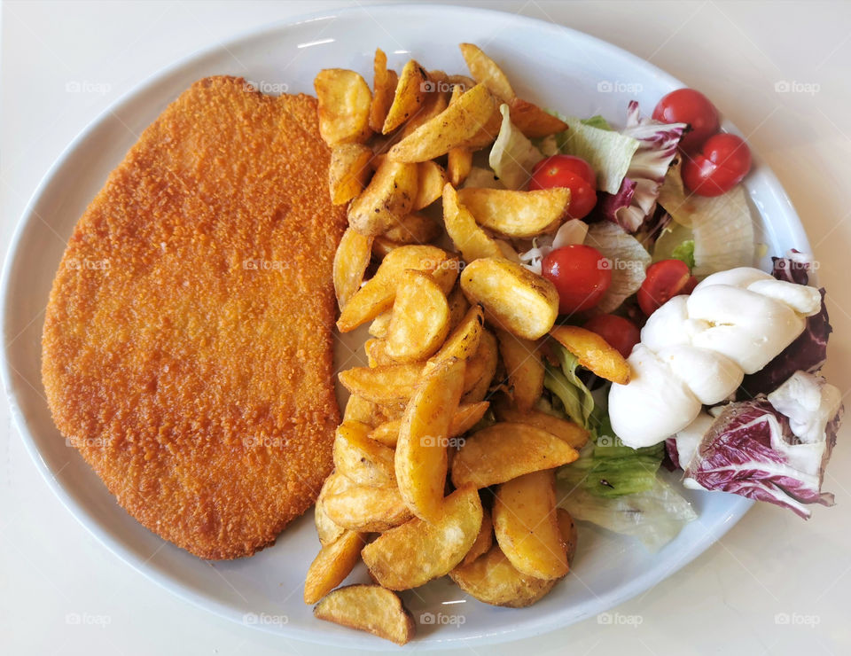 Chicken Escalope with fried potatoes or chips, vegetable salad (tomatoes, lectuce, and purple cabbage) and white egg.
