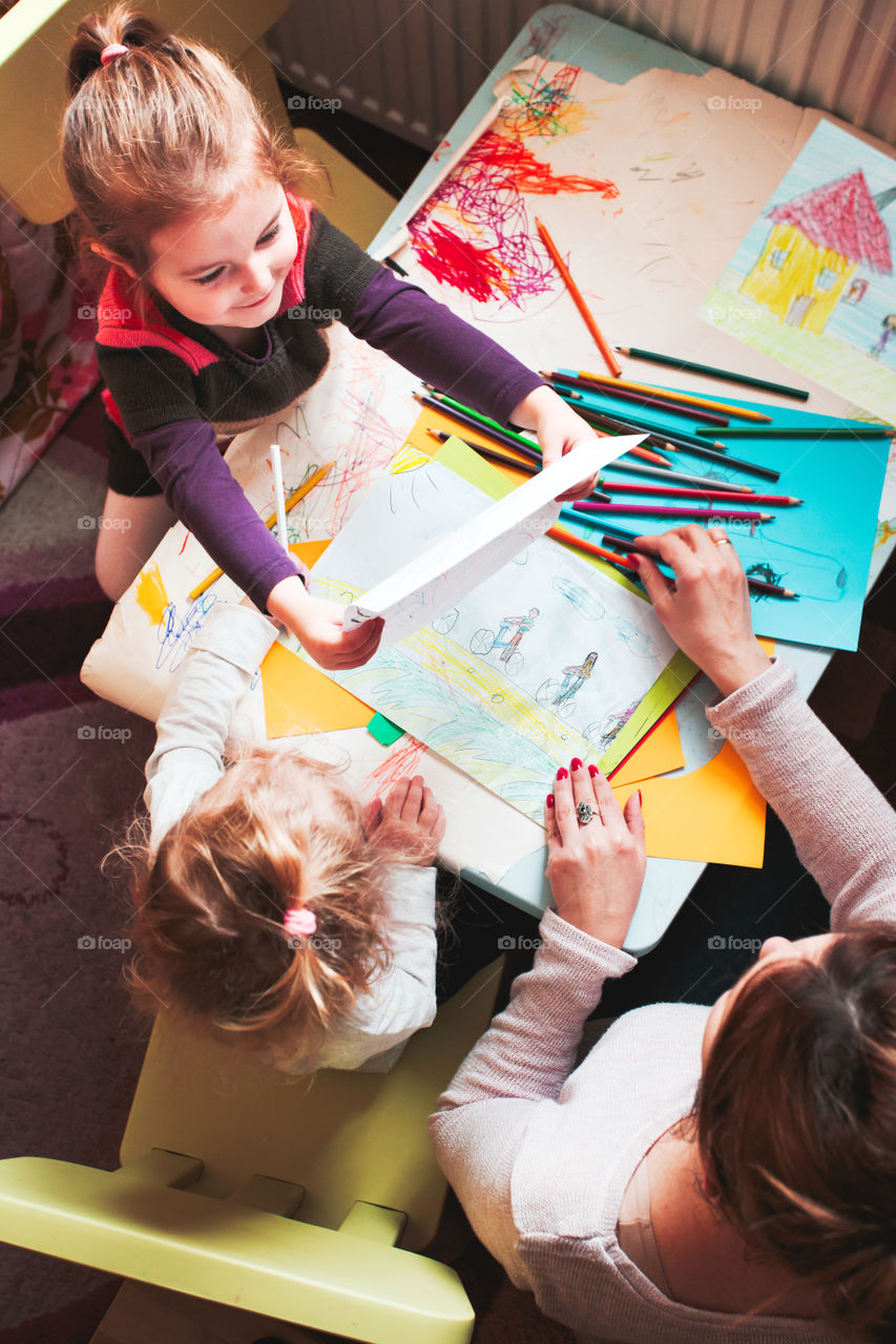 Little girl showing her picture her mom while she teaching children drawing a colorful pictures using pencil crayons sitting at table. Photo from above