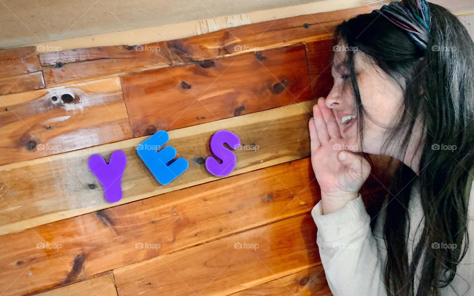 The Word Yes Written With Purple And Blue Foam Letterings On Wooden Wall As If It Is Coming Out Of The Woman’s Mouth With Happiness 