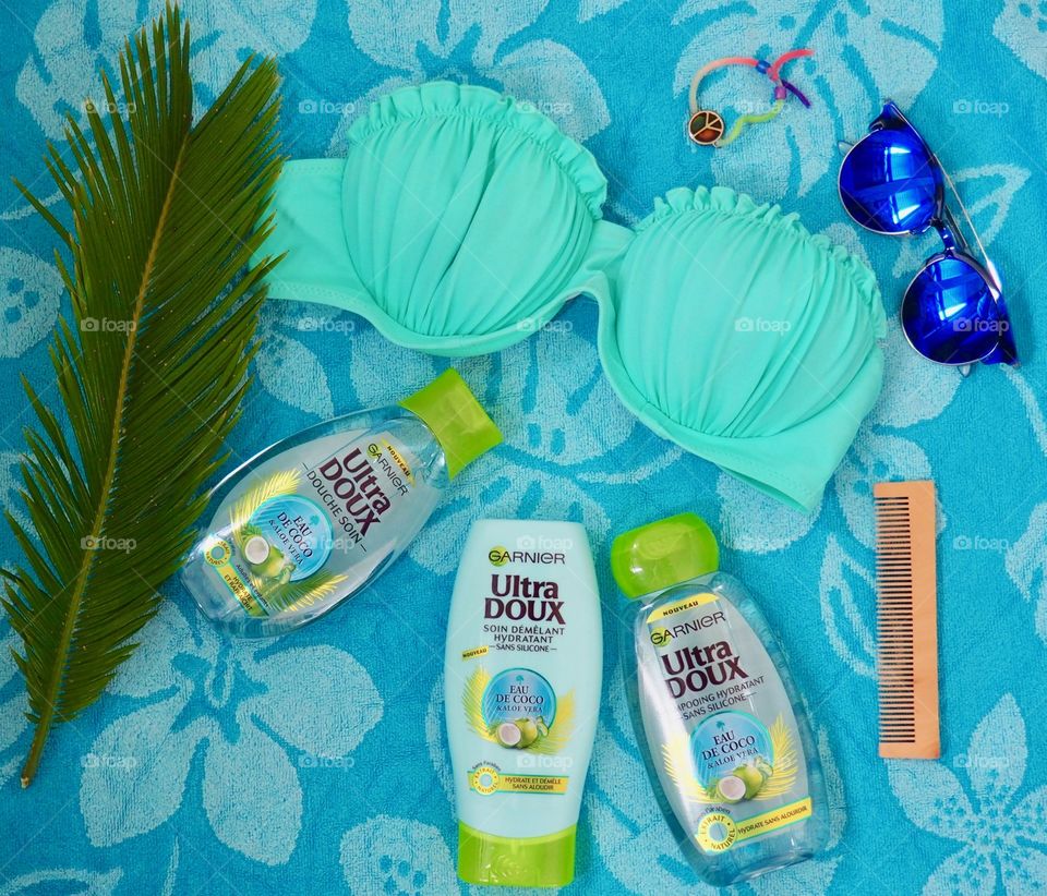 Garnier eau de coco shampoo, conditioner and bath gel on a blue towel with comb and palm leaf and blue sunglasses and bikini.