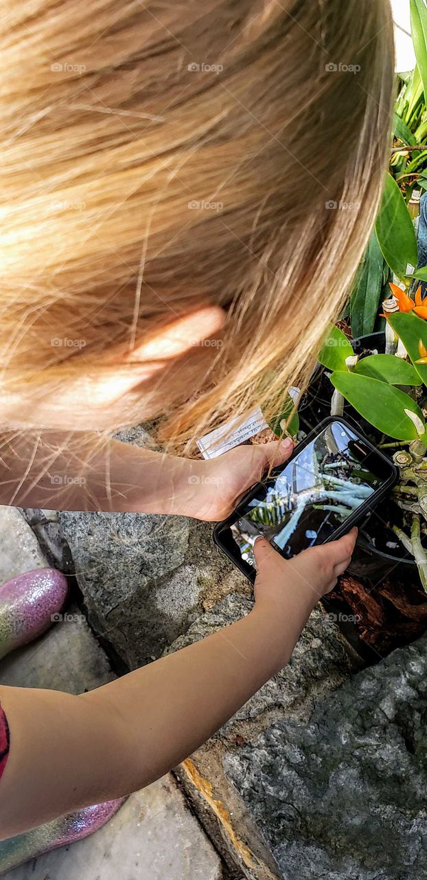 Young strawberry blonde girl taking a photograph of plant life using her camera at the park.