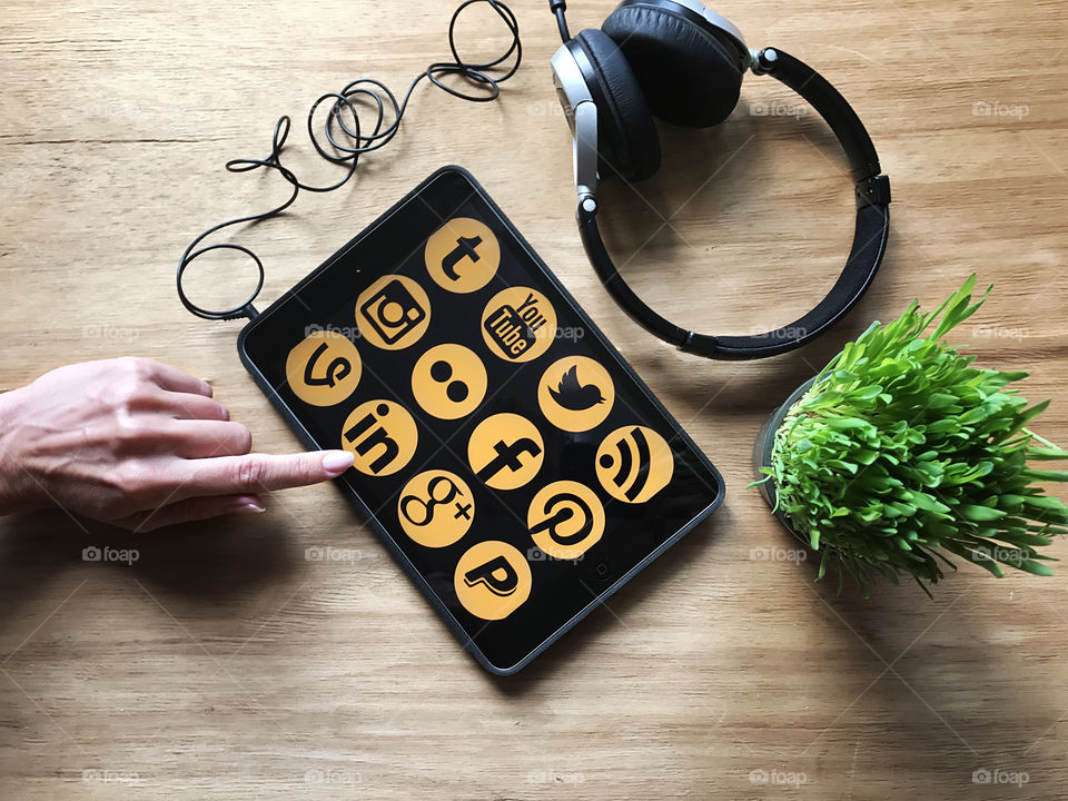 Female hand using social media on the tablet on wooden table with headphones and green house plant 