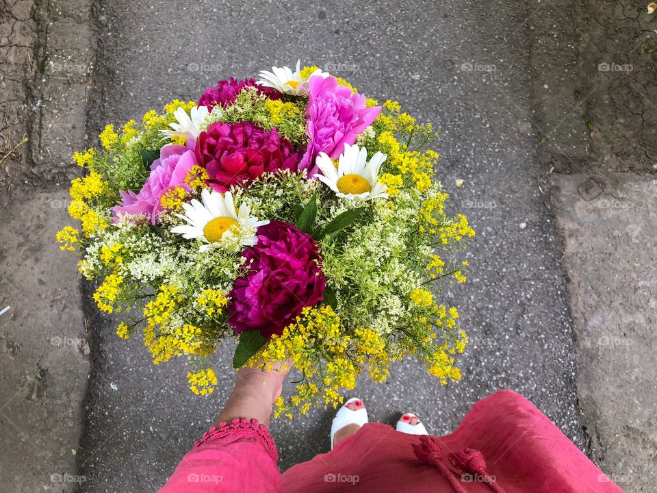 Woman in pink dress holding a giant bouquet of summer flowers consisting of pink peonies, daisies and yellow goldenrod