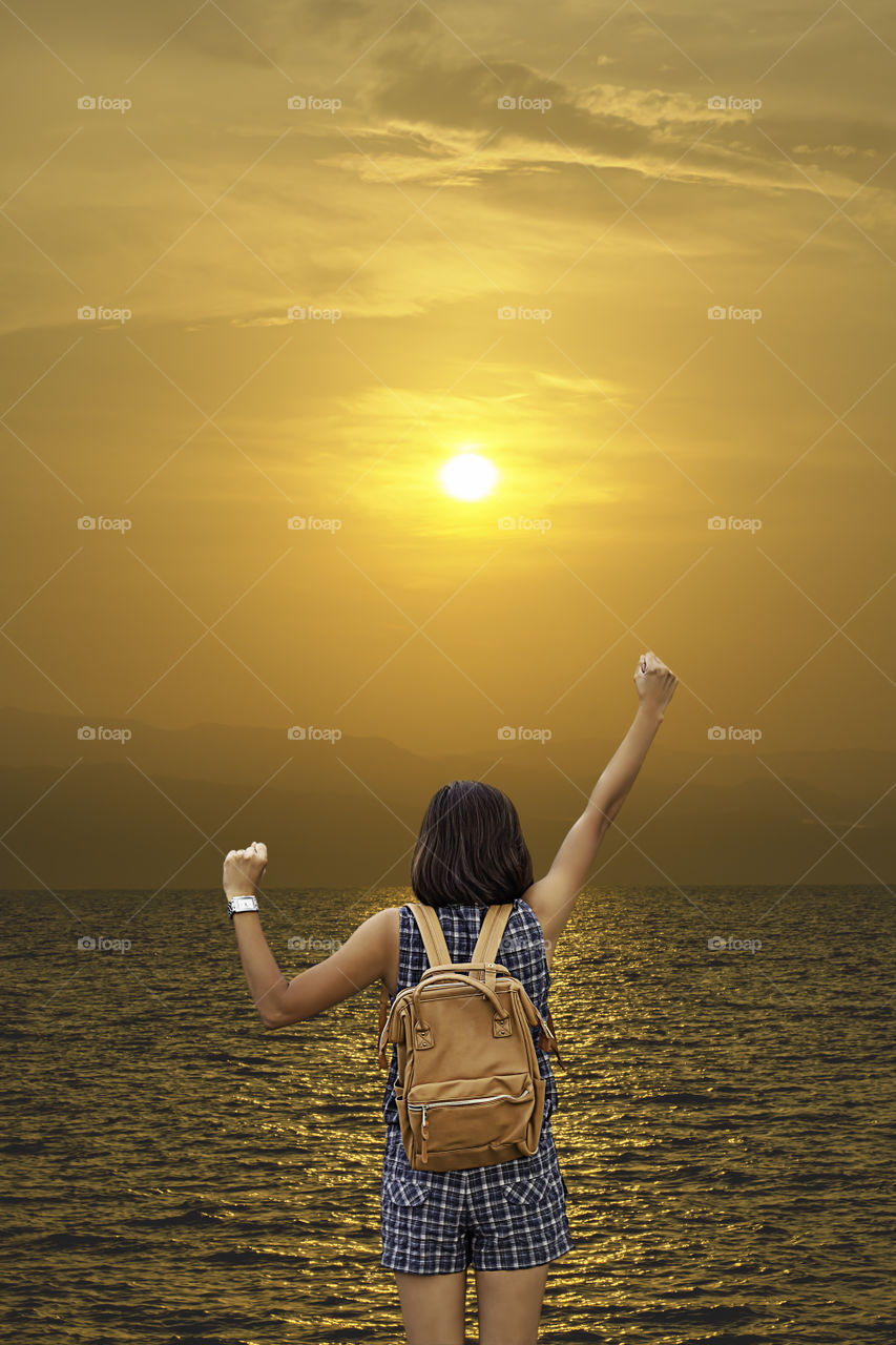 The image behind the woman raise their arms and shoulder backpack Background Golden light of sunrise behind the mountains in the sea.