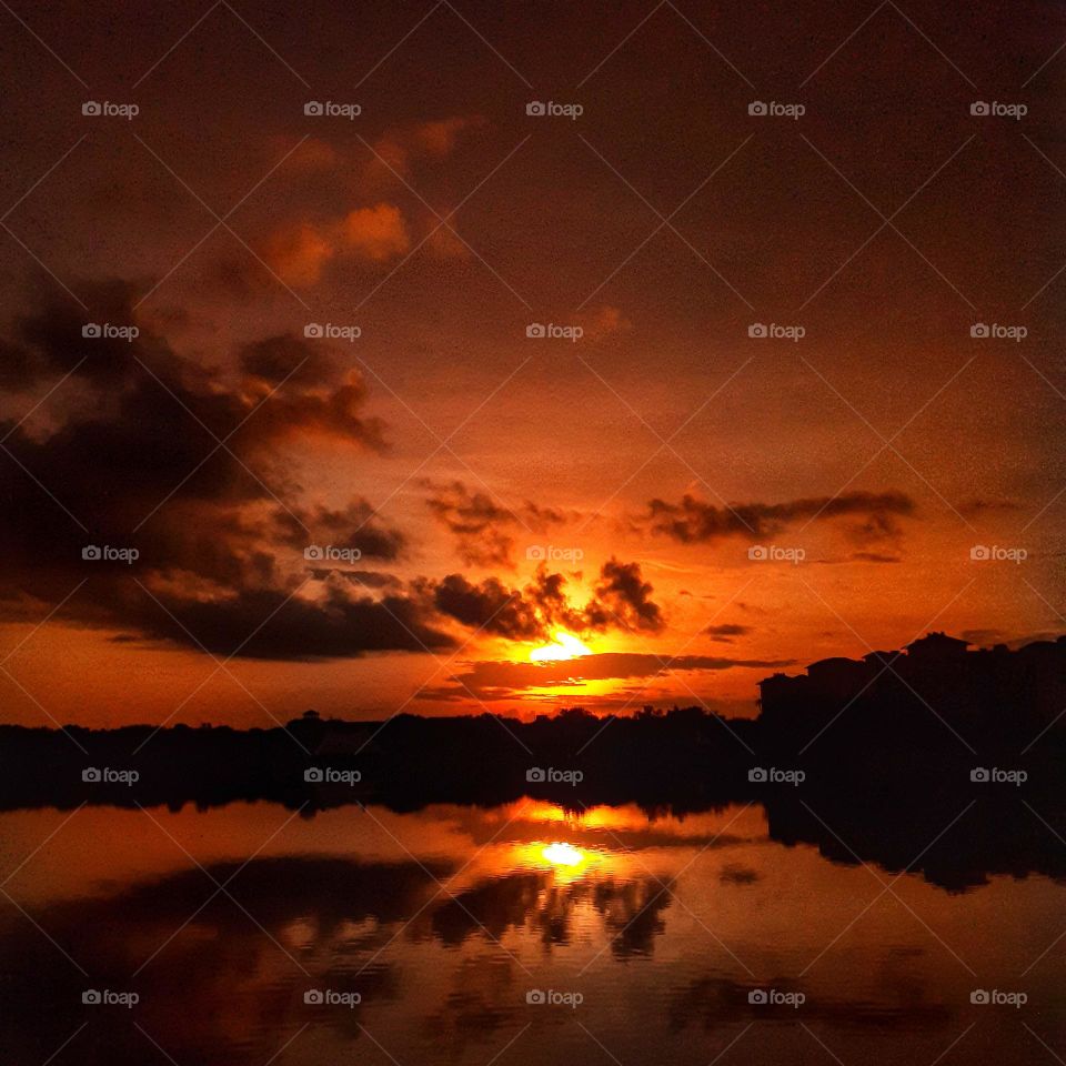 An orange and yellow sunrise and dark clouds is reflected in the water of the lake at Cranes Roost Park in Altamonte Springs, Florida.