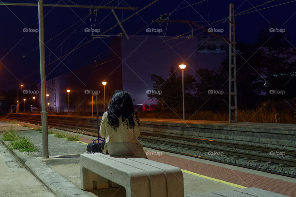A young lonely woman with a suitcase is waiting for the train arrival on an empty train station at night in Spain.