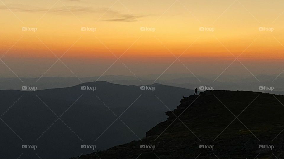 Man looking at sunrise. Mountains at sunrise. Man standing on peak. Natural mountain landscape with illuminated misty peaks, foggy slopes and valleys, blue sky with orange yellow sunlight. Amazing scene from Beskid Zywiecki in Poland