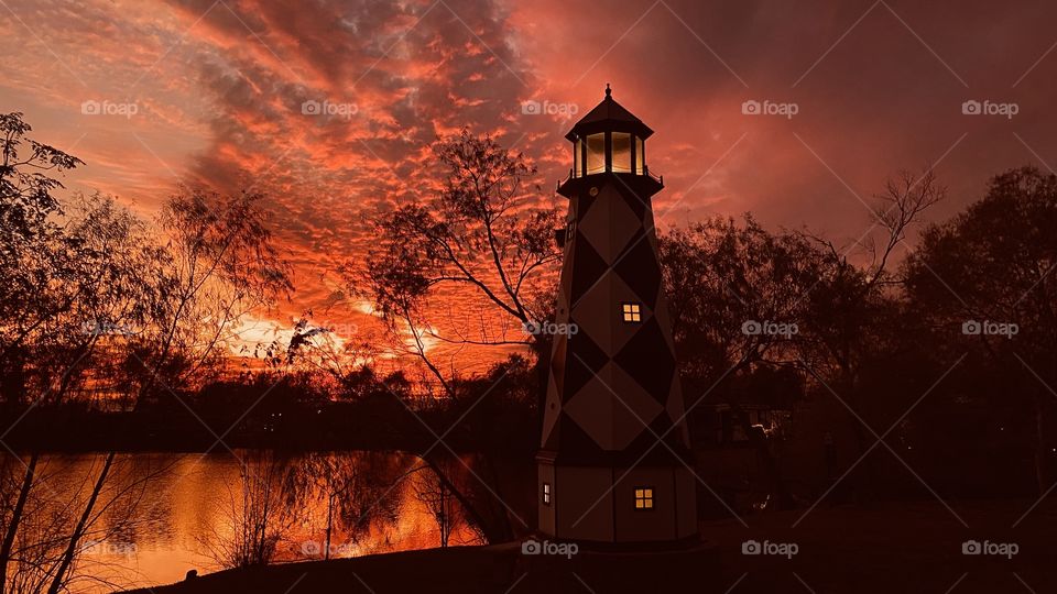 LightHouse Setting in s Gorgeous Sunset-Twilight Bright Fire Orange mix with Deep Dark Clouds grouped tightly Together. Reflections if Fiery Orange Mix can seen on Lake Water. Beautiful Scenery.