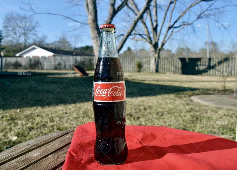 A glass bottle of Coca-Cola on a wooden table top with a red linen outdoors in a backyard