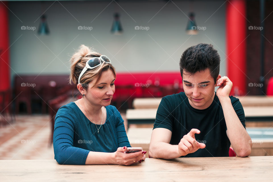 Woman and young man talking together and using mobile phones sitting by a table in cafe