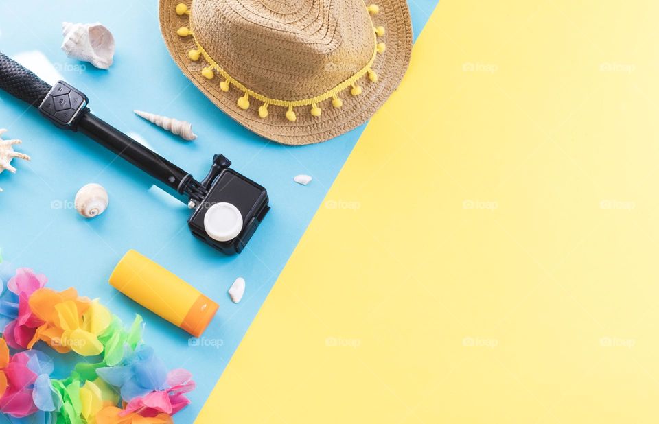 A straw hat, camera, sunblock, seashells and a coas flower wreath lie on the left on a blue-yellow ion with copy space on the right, close-up of a flat lay. Concept summer, blogger, travel.