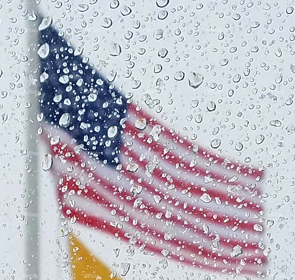 American Flag 🇺🇸 seen through rain droplets on window. Flag is waving in rain from top of flag pole.