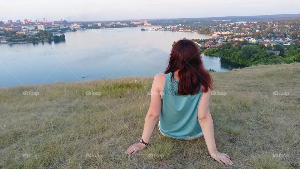 girl looks into the distance, meditation, contemplation nature, view from the mountain to the city, pond, water, summer, evening, calmness, peace, light breeze, the girl is sitting on the grass