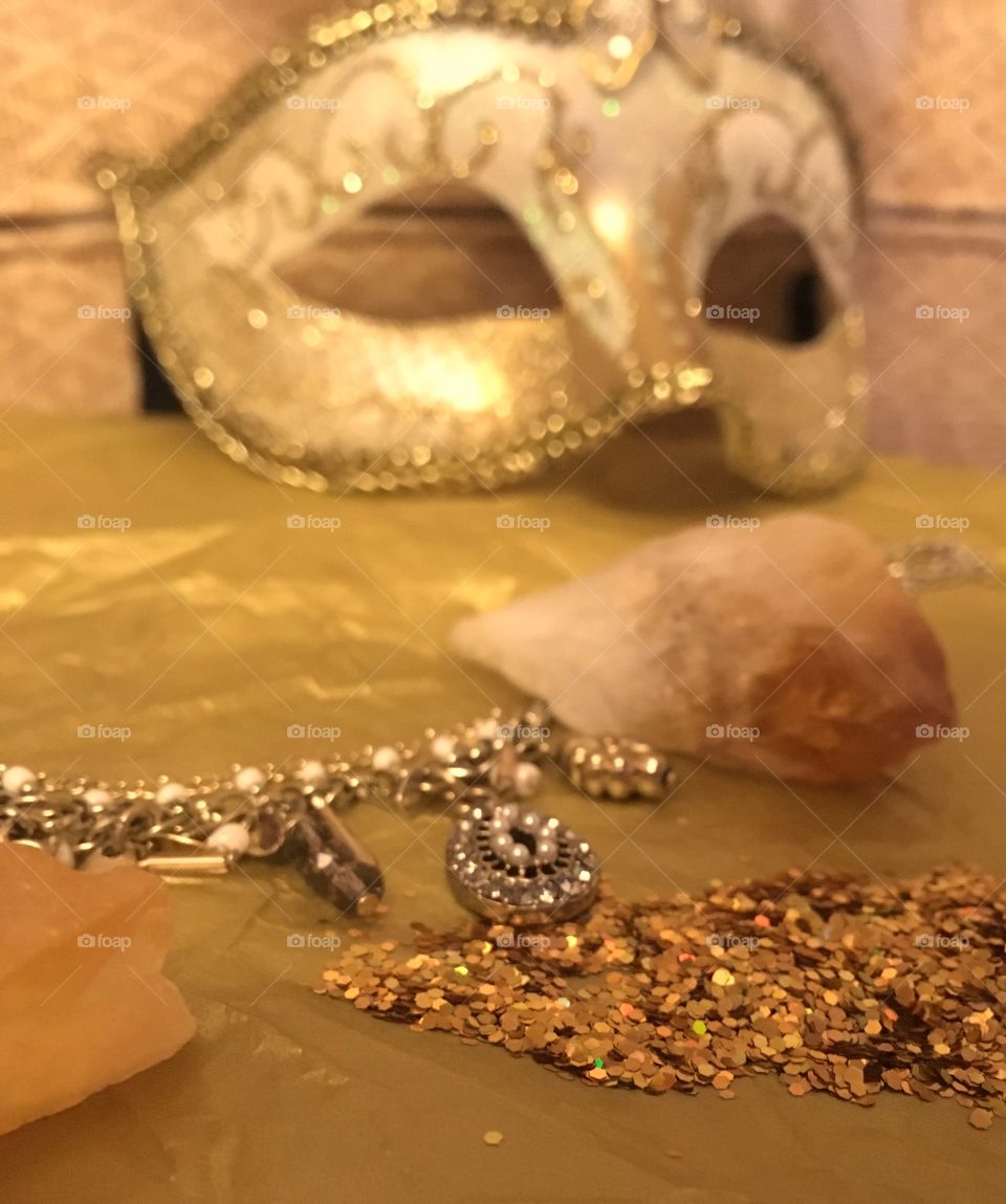 Gold glitter spilled with a necklace and calcite stone on the left, citrine crystal rock on the right and Venetian masquerade mask in the background 