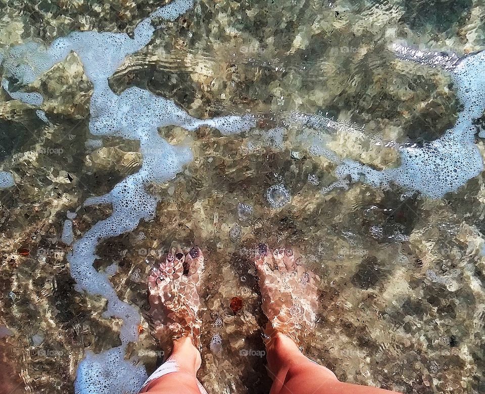 a girls feet in the shallow water of an incoming wave with seashells and foam framing her painted toes