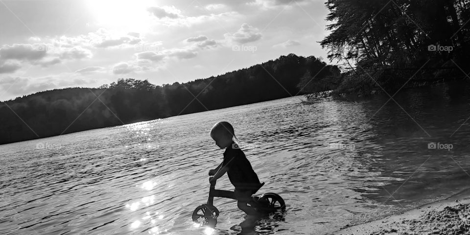 Toddler boy rides his bicycle into the lake at sunset. silhouette.