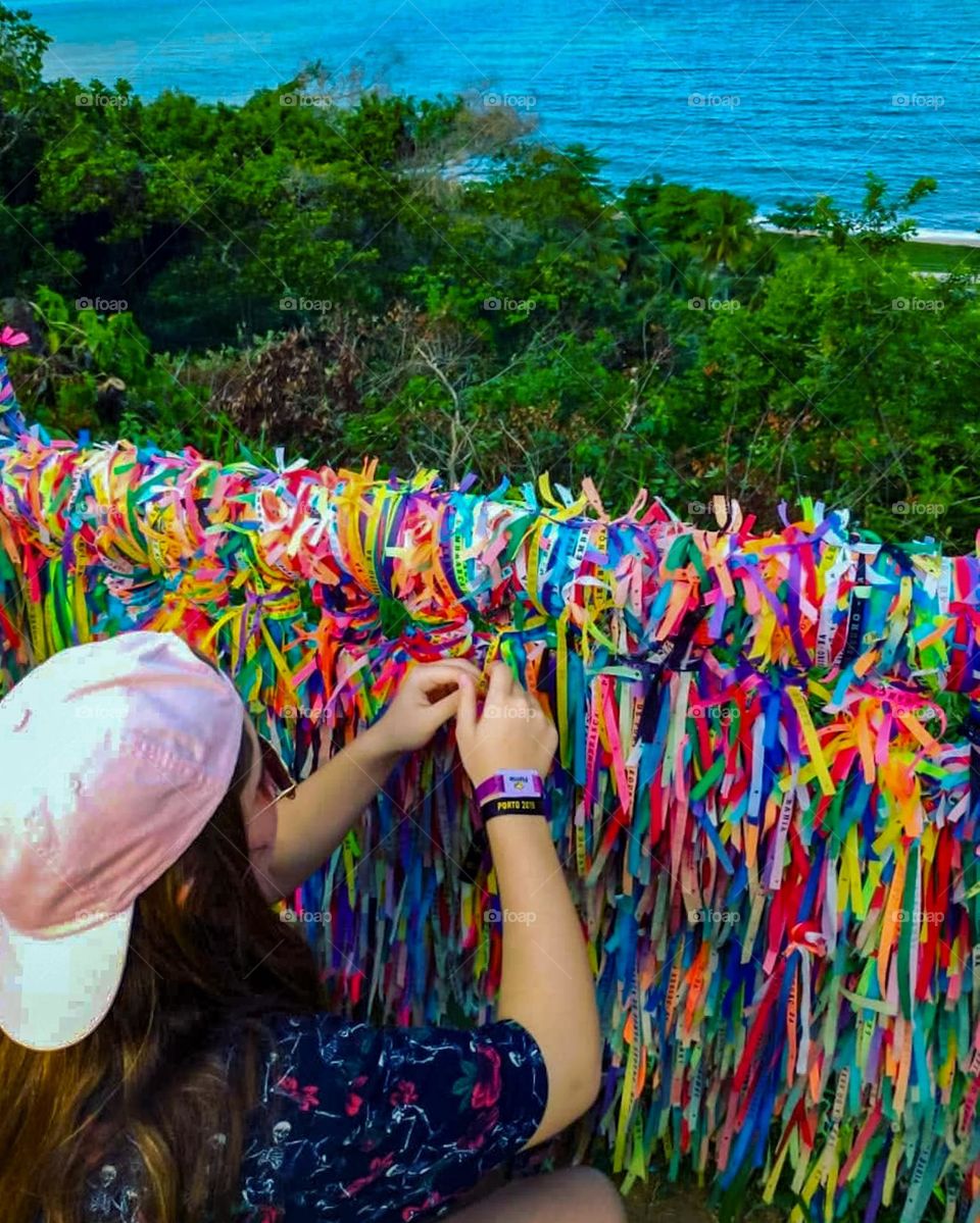 Teenager ties the ribbon of Senhor de Bomfim to the railing next to the Church.  Superstition says that Bonfim's ribbons of different colors carry the power to fulfill three wishes. Porto Seguro Beach, Bahia Brazil, in the background.