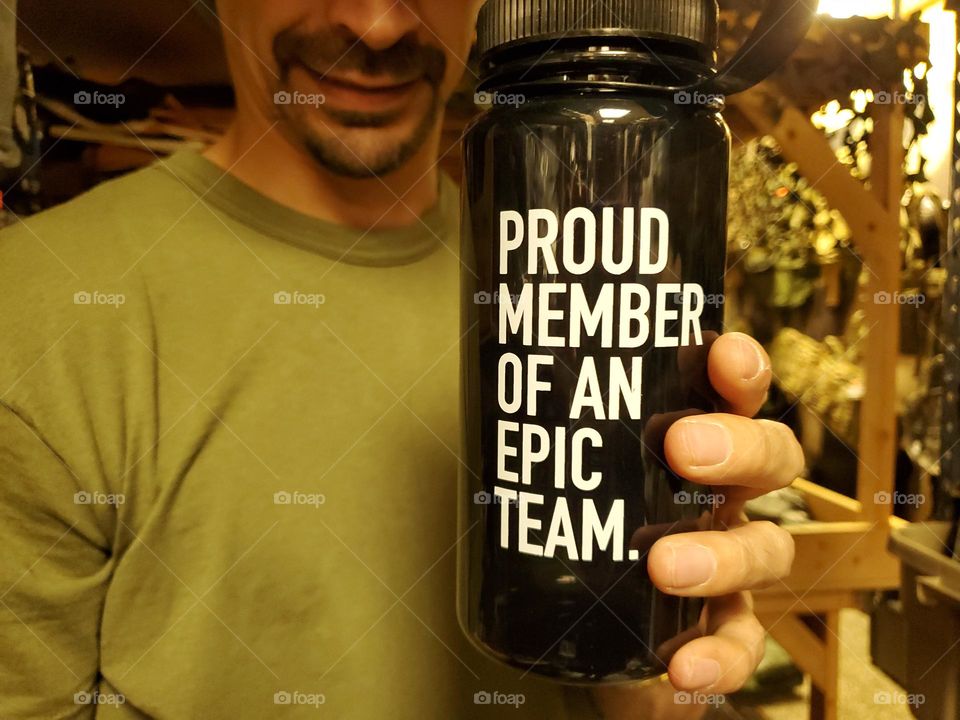 A man holding a black water bottle with the message: proud member of an epic team.