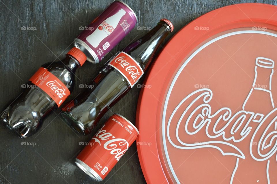 A Coca-Cola flat lay with a can of Coke a can of cherry Coke a glass bottle of Coca-Cola and a plastic bottle of Coca-Cola with a black background