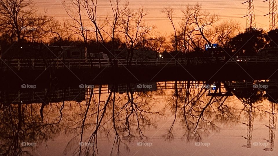 Sunset. Winter. Winterized trees on Landscape. Horizon full of Colors during Twilight reflections of trees, Branches, limbs, twigs. Reflections of the Shore Landscape fences, buildings, trees.