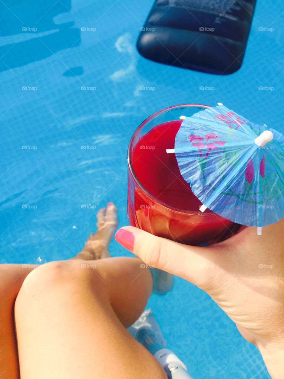 Woman hand with pink nail polish holding glass of raspberry red juice with blue umbrella over the pool blue water