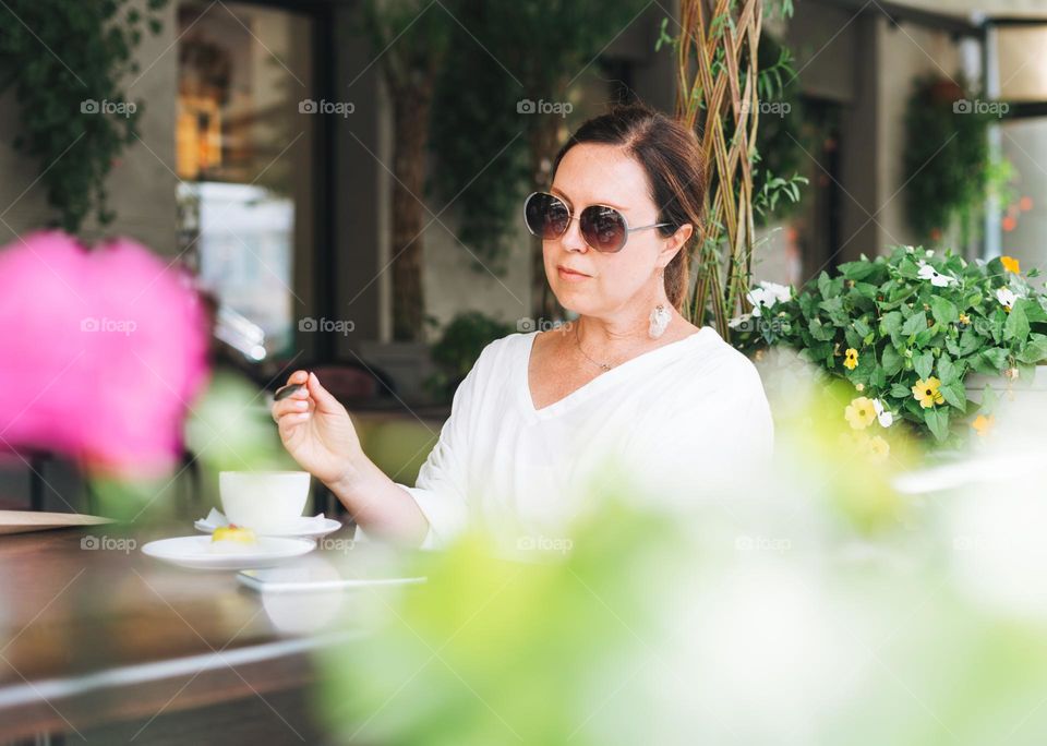 Portrait of smiling beautiful brunette middle aged woman in sunglasses in white clothes with coffee at terrace of the summer cafe