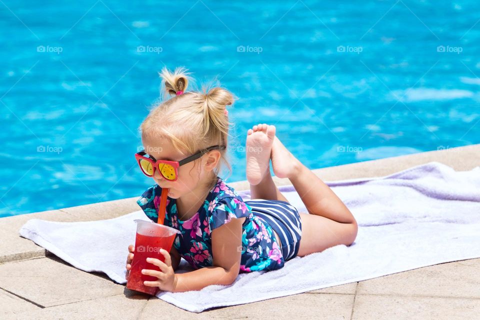 Happy little Caucasian girl drinking fresh juice near swimming pool at hot summer day 