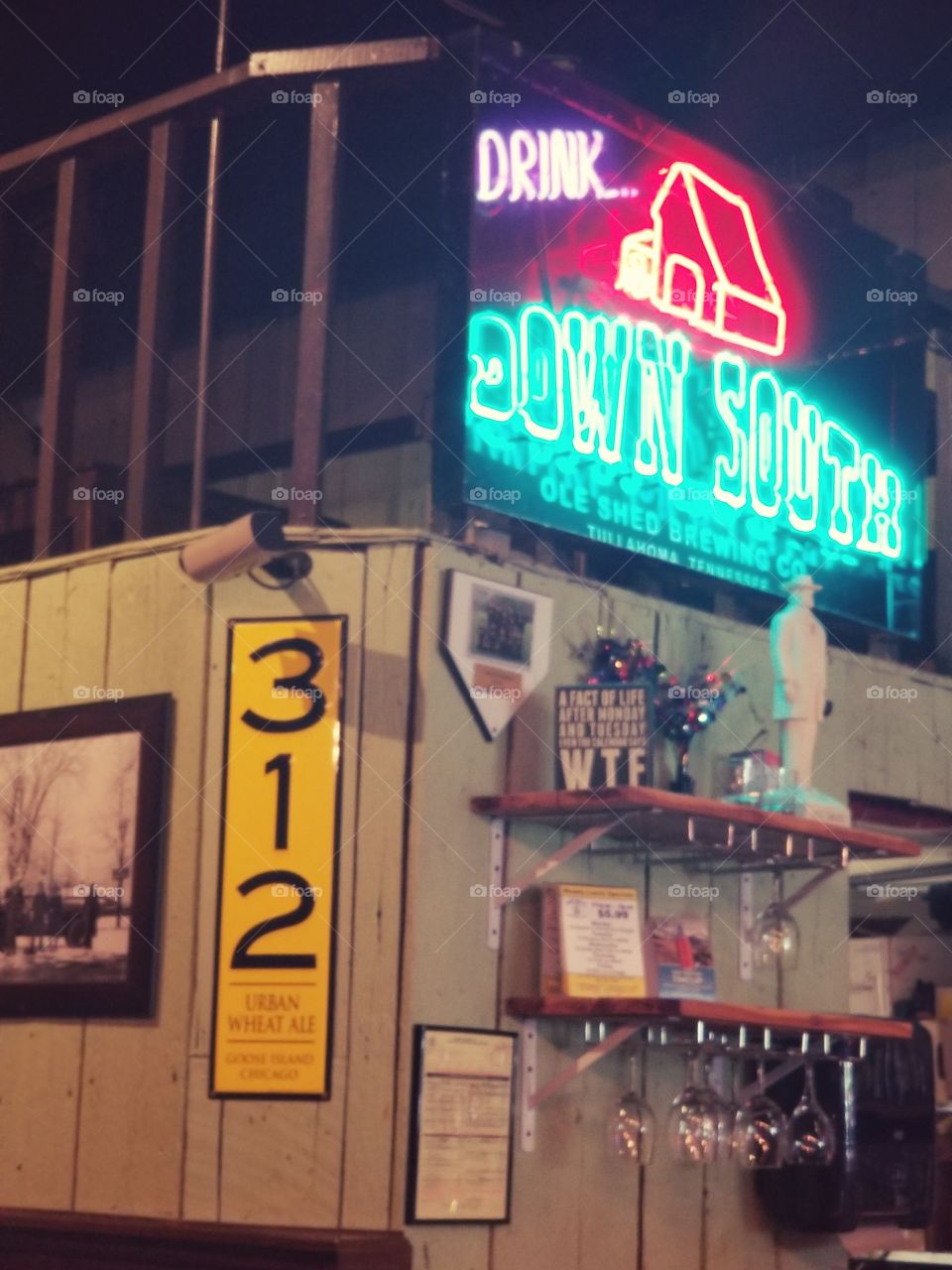 Neon glow of colorful sign promoting Down South atmosphere, lights up inside of typical Americana bar scene speckled with vintage signage 312, glasses, and pictures.