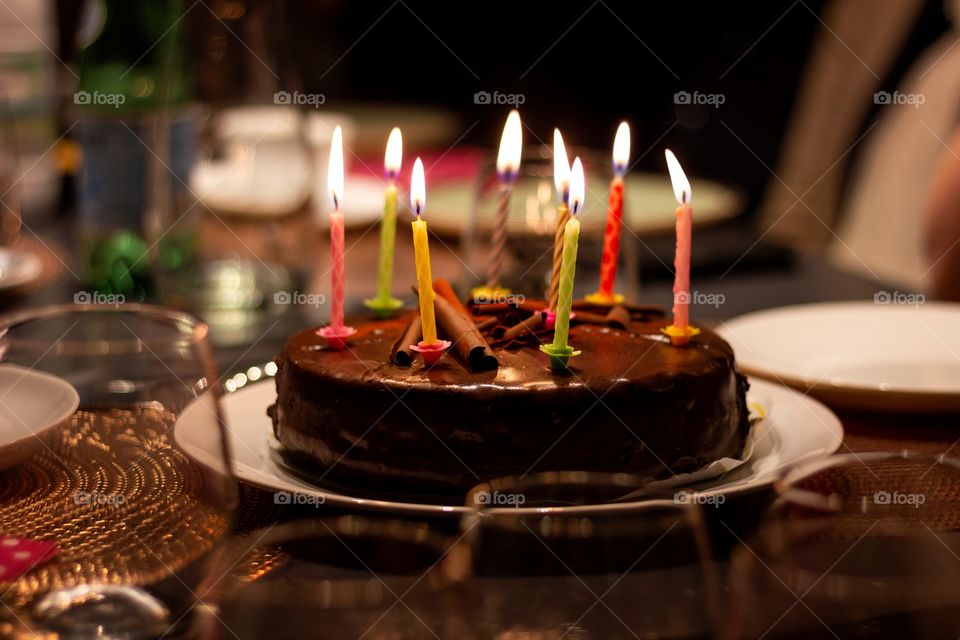 A portrait of a birthday cake with lit candles on it. the cake is chocolate flavoured and all candles are differently colored.