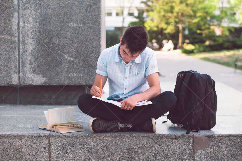 Student making the notes learning from books sitting on a monument outside of university. Young boy wearing a blue shirt and dark jeans