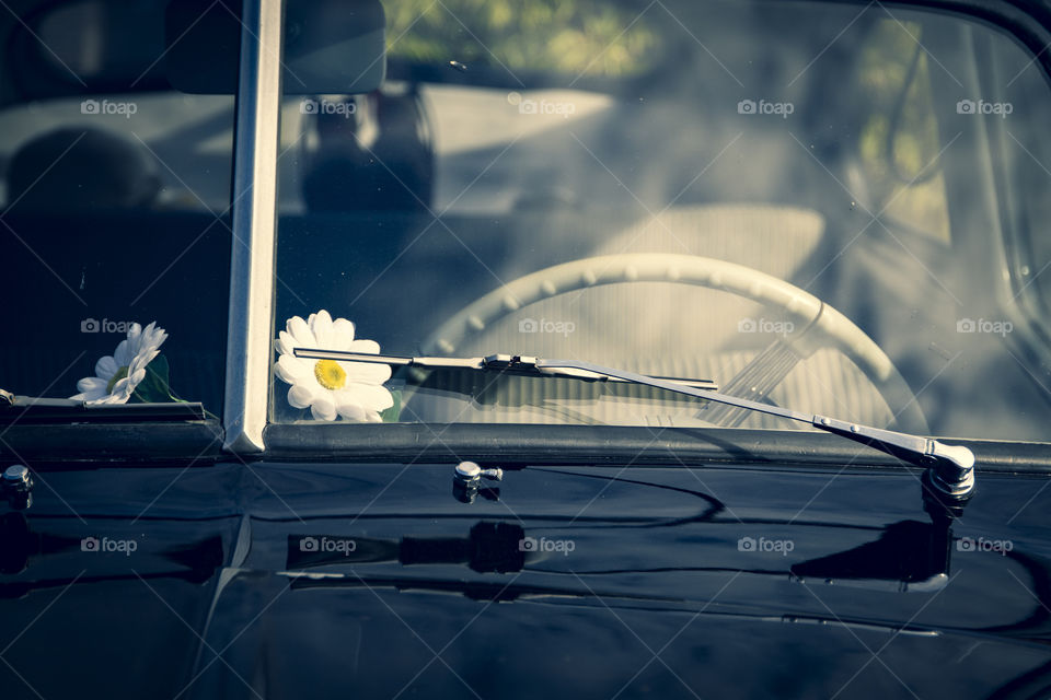 A portrait of the wind shield of a classic car with a flower on the dashboard. hou can see the steering Wheel and the wind shield wiper.