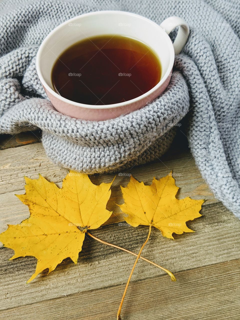 A cup of hot tea wrapped in a sweater and yellow maple leaves on a wooden board