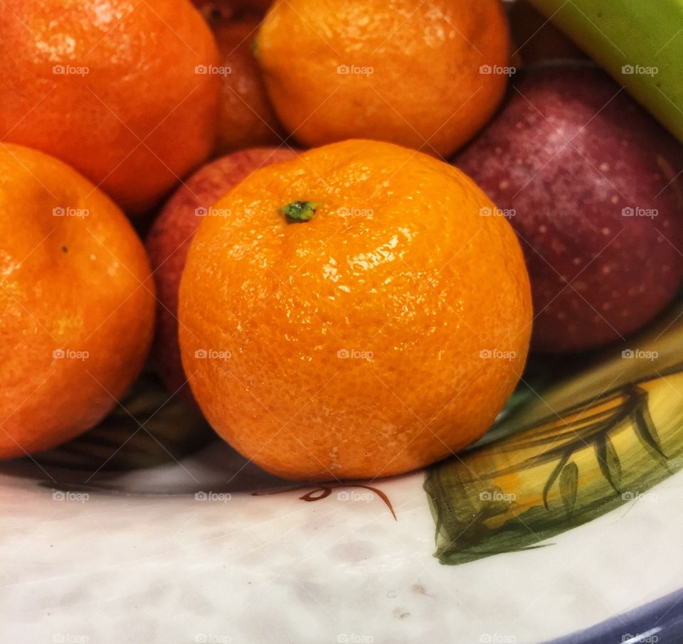 A clementine, a kind of small orange, sits in a decorative bowl with other fruit. This is a close shot.