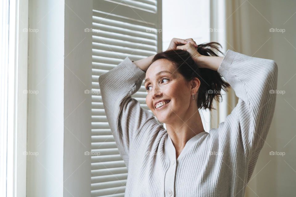 Young beautiful woman forty year with brunette hair in cozy knitted cardigan near window in bright interior at home