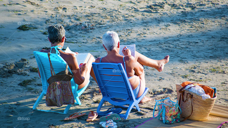 Two senior women relaxing at the beach during summertime solving crosswords and reading