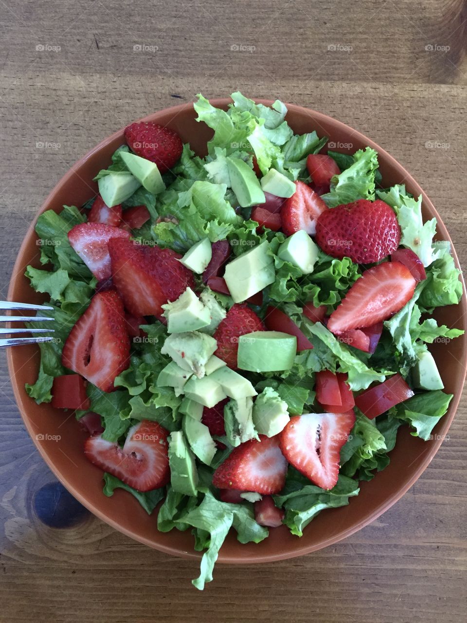 Circular ceramic orange bowl with healthy salad of leaf lettuce, strawberries, avocados, dressing with fork on kitchen table ready for lunch, dinner, or snack