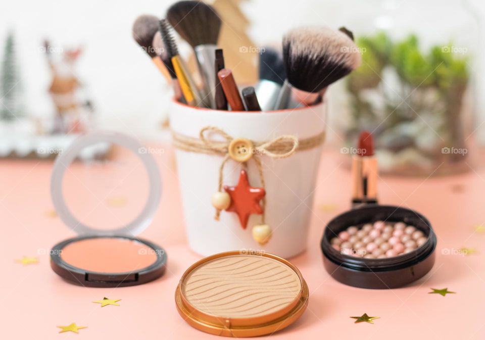 Set of natural cosmetics of foundation face powder, red lipstick, brushes with eye pencils in a festive bucket standing on a pink background with blurry holiday decorations and white brick wall,side view. Concept female cosmetics,beauty salon.