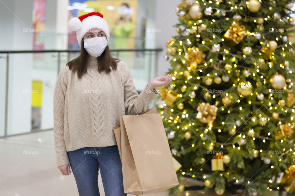 Happy woman in a santa claus hat and a cozy sweater in a shopping center near the christmas tree Christmas shopping wearing a facemask. Concept: holiday shopping during the coronavirus epidemic a woman wearing a medical mask