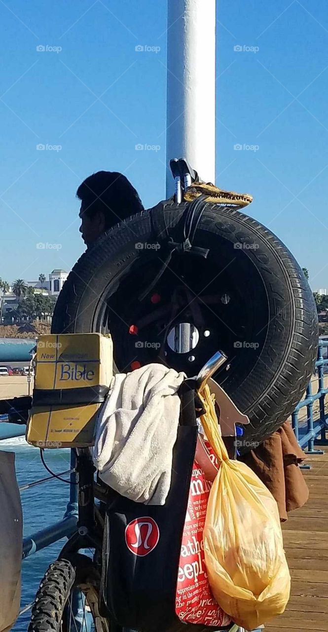 Man on a bicycle with a auto tire, an alligator head held together with a cord and a Bible on the back while taking in the Pacific Ocean on the Santa Monica pier