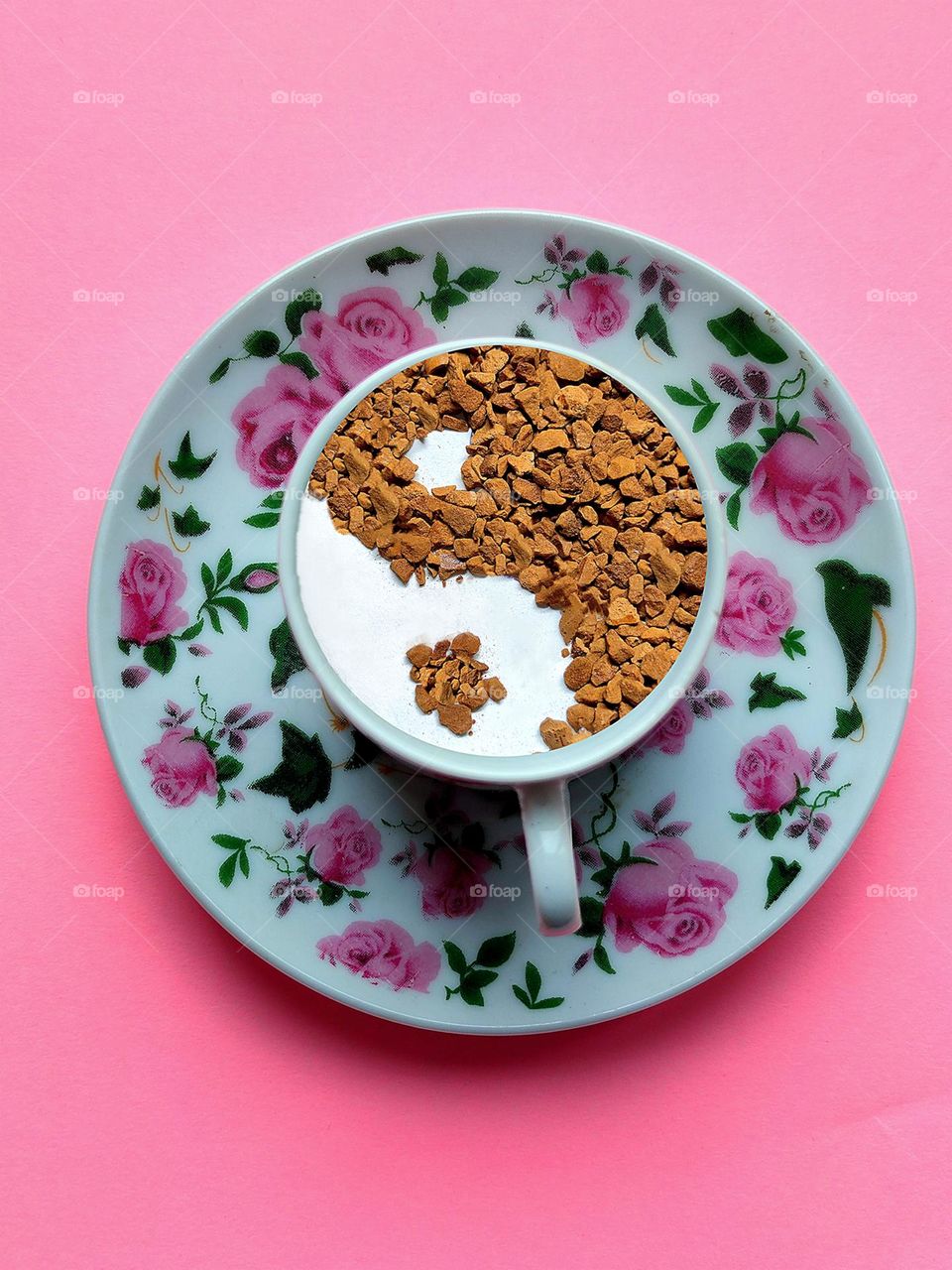 View from above.  On a pink background is a saucer with pink flowers, on which stands a coffee cup.  The cup contains white milk and coffee granules in the pattern "Yin and Yang", "Black and White". Philosophy and coffee