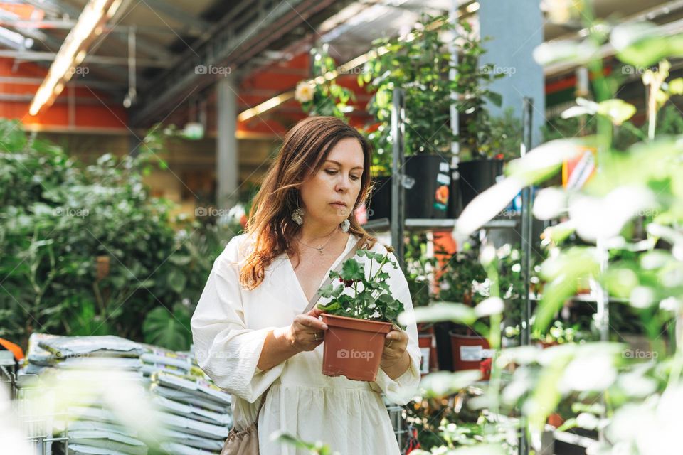 Brunette middle aged woman in white dress buys green potted house plants at the garden store