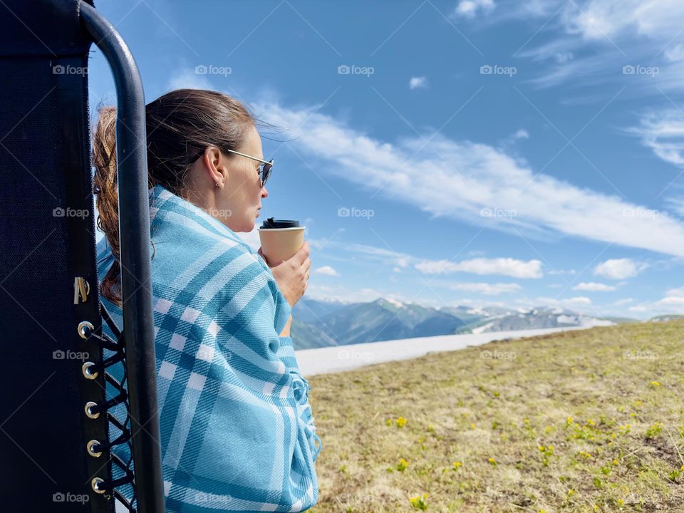 girl in a blanket with a cup of coffee sits in a chair in the mountains