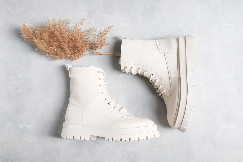 One pair of new white leather martin boots with a beige dried flower branch lie on a light gray cement background, flat lay close-up.