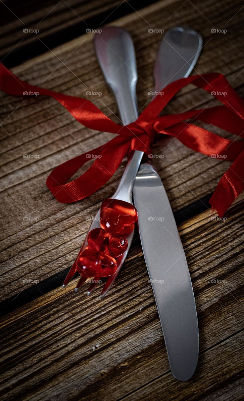 A beautiful view of a fork and knife tied with red ribbon and three glass hearts lying on a brown wooden table, side view.