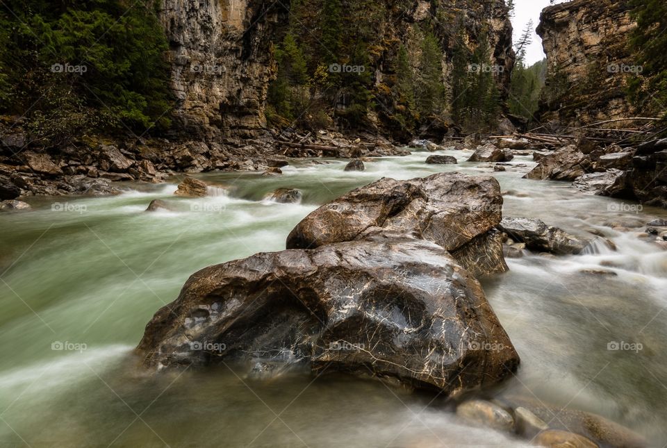 Large boulders sit in the green waters of Toby creek in a steep canyon with cascading water.